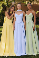 Three women standing outdoors wearing pastel floor-length corset gowns — one in light yellow with a flowing scarf detail, one in baby blue with a halter neck strap, and one in sage green with thin straps — posing in a garden setting