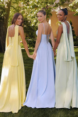 Three women standing outdoors wearing pastel floor-length corset gowns — one in light yellow with a flowing scarf detail, one in baby blue with a halter neck strap, and one in sage green with thin straps — posing in a garden setting