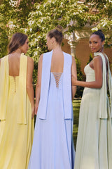Three women standing outdoors wearing pastel floor-length corset gowns — one in light yellow with a flowing scarf detail, one in baby blue with a halter neck strap, and one in sage green with thin straps — posing in a garden setting