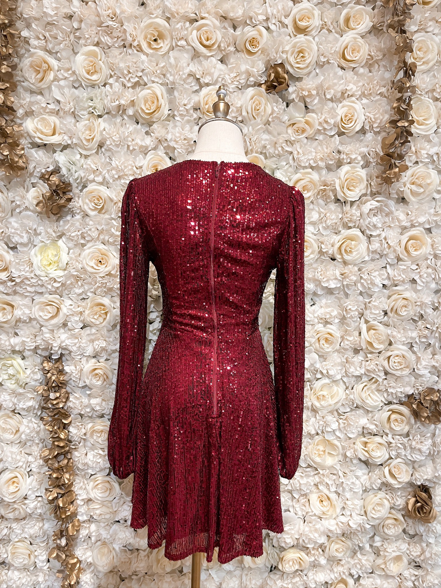Burgundy sequin mini dress with cutouts and puff sleeves, displayed on a mannequin against a floral backdrop.