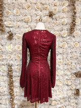 Burgundy sequin mini dress with cutouts and puff sleeves, displayed on a mannequin against a floral backdrop.