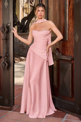 A woman models a floor-length dusty pink halter gown with a draped, asymmetrical wrap-style skirt, standing in an open dark wooden doorway.