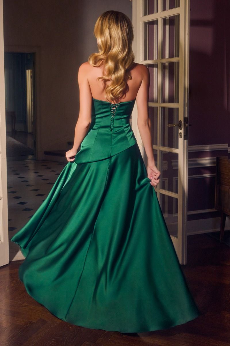 A woman viewed from behind wearing a strapless emerald green satin ball gown with a corseted back and full skirt, standing in an indoor room with wooden floors and glass-paneled doors.