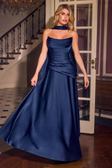 A woman modeling a floor-length navy blue satin gown with a halter-style neckline and draped waist details, standing in an indoor hallway with a staircase and glass doors in the background.