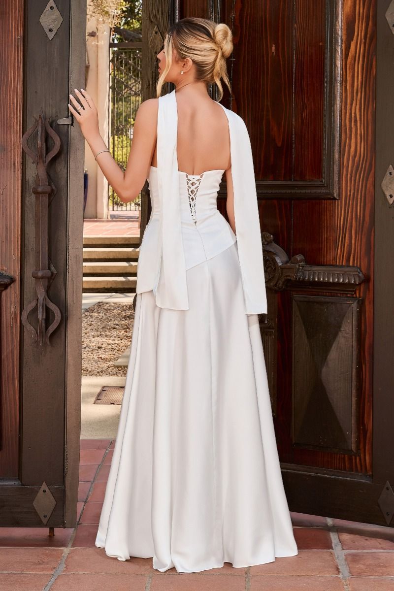 A woman stands with her back to the camera wearing a white sleeveless gown with a draped cape-like overlay and lace-up open back, posed in front of dark wooden doors.