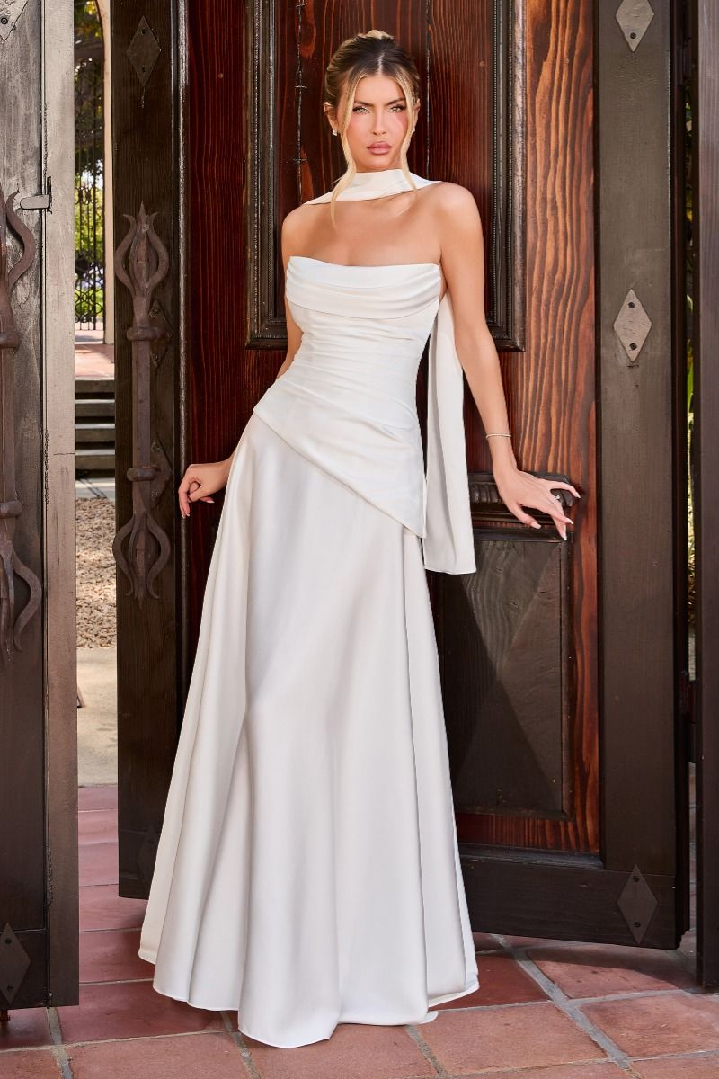 A woman models a floor-length white evening gown with a halter-style neckline, ruched bodice, draped side detail, and a full skirt, standing in front of dark wooden doors.