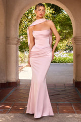 A woman models a light pink one-shoulder mermaid gown in a stone archway garden setting.