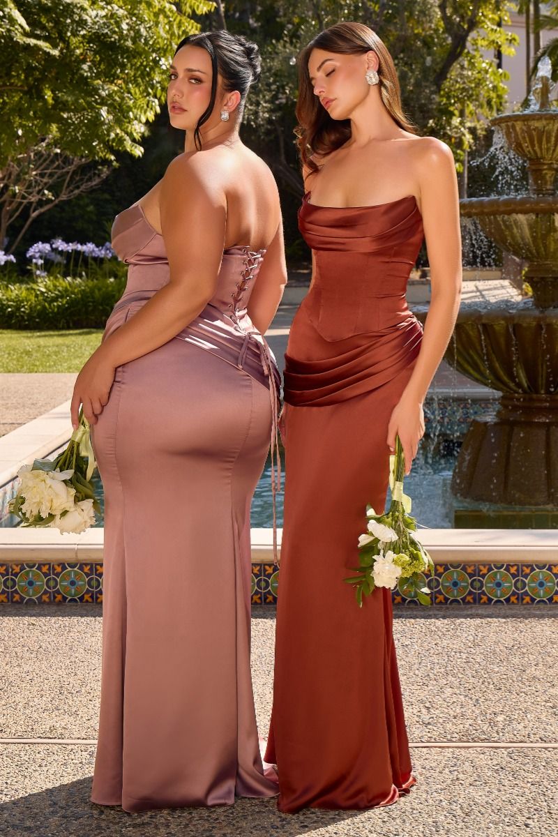 Two women modeling floor-length strapless bridesmaid dresses outdoors beside a fountain. The woman on the left wears a dusty mauve satin gown and faces away, while the woman on the right wears a rust-brown satin gown and faces forward, holding white flowers.