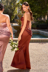 A woman wearing a strapless rust-brown satin mermaid gown stands outdoors beside a pool with a fountain in the background. Another woman in a matching dusty rose gown is partially visible at the left edge, and the main model holds a bouquet of white flowers.