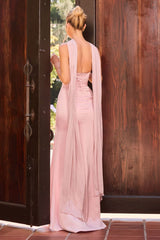 A woman stands with her back to the camera wearing a floor-length blush pink gown with a draped open back, posed between two dark wooden doors.