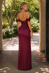A woman stands in a garden archway wearing a floor-length off-the-shoulder gown from the back. The dress is a rich deep burgundy with a satin-like sheen and structured back detail.