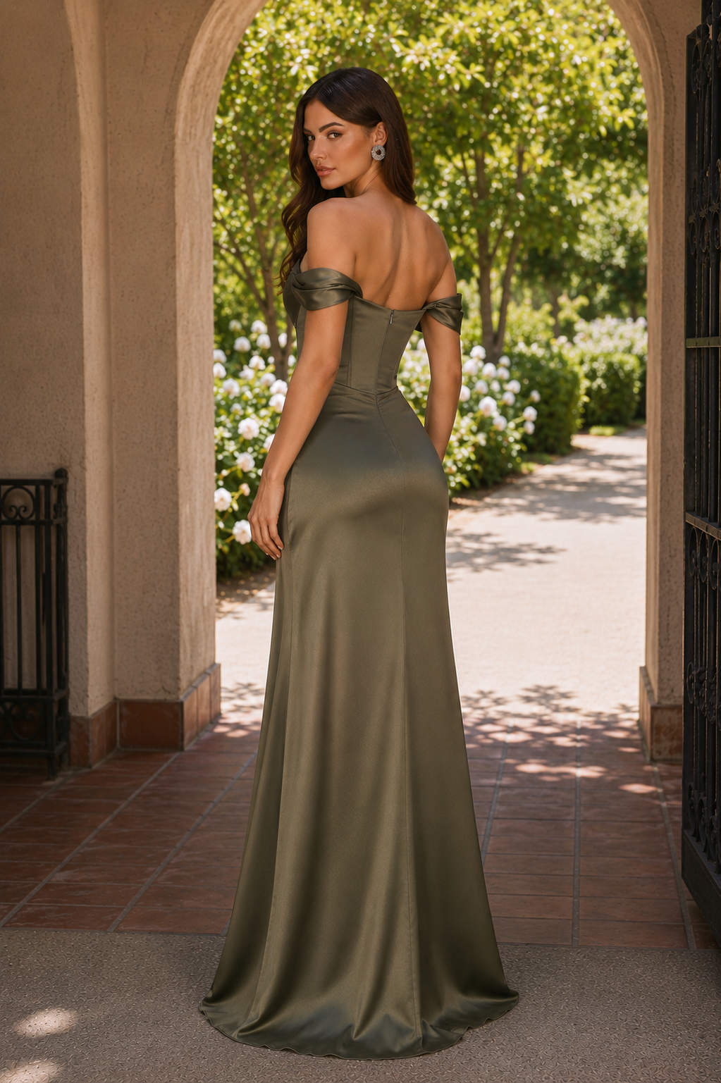 A woman stands in an outdoor archway wearing a floor-length off-the-shoulder satin gown in an olive green color. The dress features a fitted corset-style bodice and a flowing skirt.