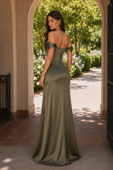 A woman stands in an outdoor archway wearing a floor-length off-the-shoulder satin gown in an olive green color. The dress features a fitted corset-style bodice and a flowing skirt.
