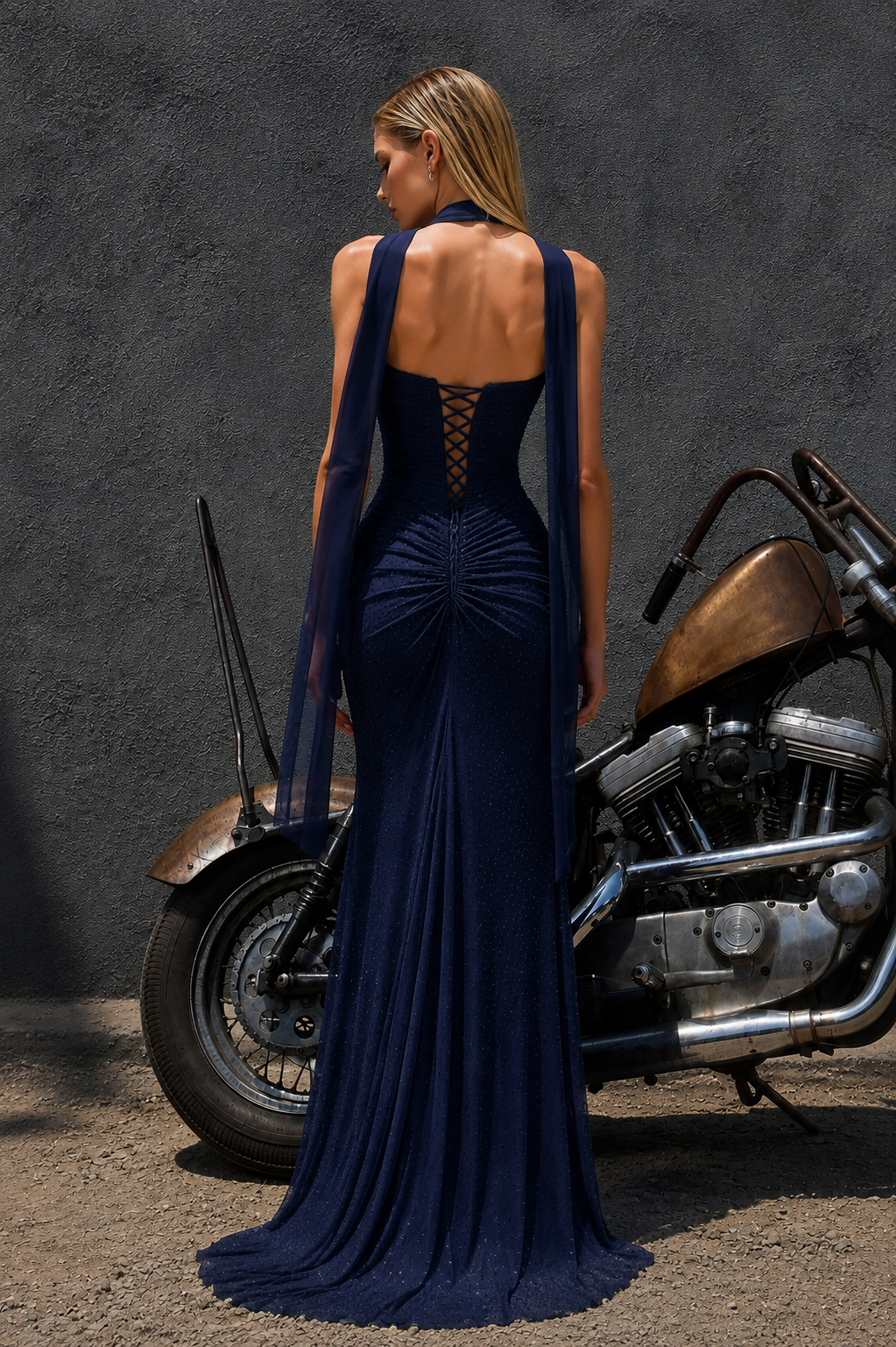 A woman stands with her back to the camera wearing a deep navy blue evening gown with a lace-up open back and sheer draped shoulder straps. She is posed outdoors next to a brown and chrome motorcycle against a dark textured wall.