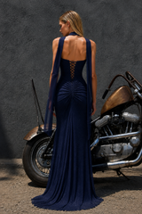 A woman stands with her back to the camera wearing a deep navy blue evening gown with a lace-up open back and sheer draped shoulder straps. She is posed outdoors next to a brown and chrome motorcycle against a dark textured wall.