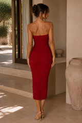 A woman wearing a strapless deep red beaded midi dress, shown from the back in a warm, sunlit indoor-outdoor setting with beige walls and stone steps.
