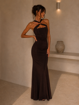 A woman modeling a floor-length, dark brown halter-neck gown with a ruched bodice and flared skirt, standing in a minimalist beige interior with a shelf and wall sconce.