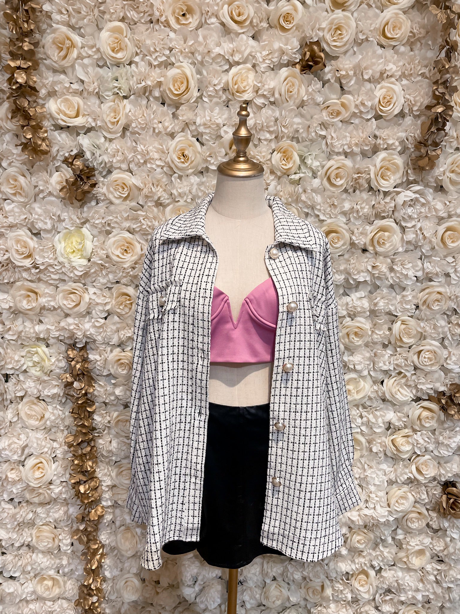 Mannequin wearing a white checkered oversized tweed shacket with a pink bra underneath, displayed in a floral-themed setting.