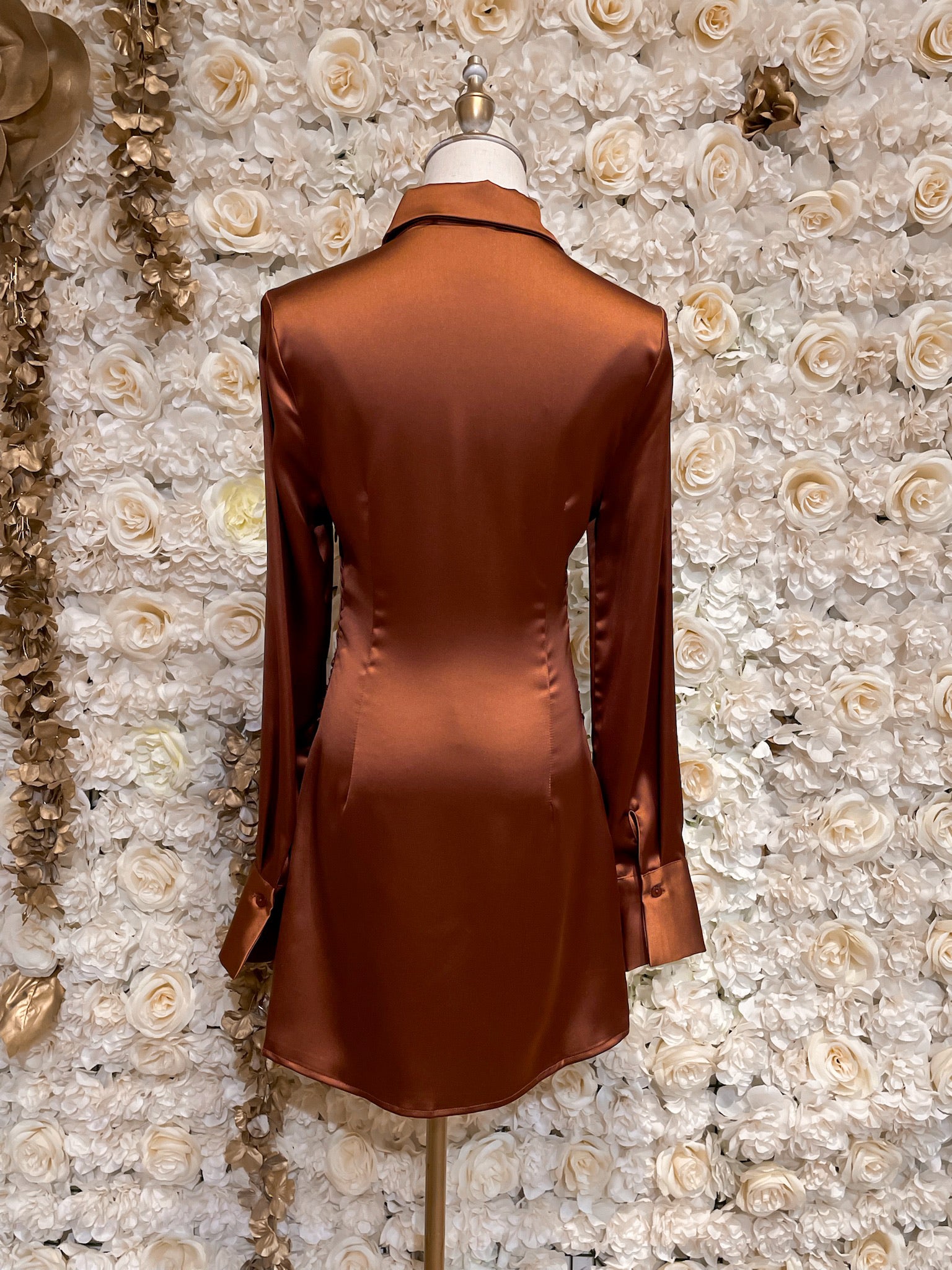 Brown satin long sleeve button up shirt dress displayed on a mannequin against a floral background.