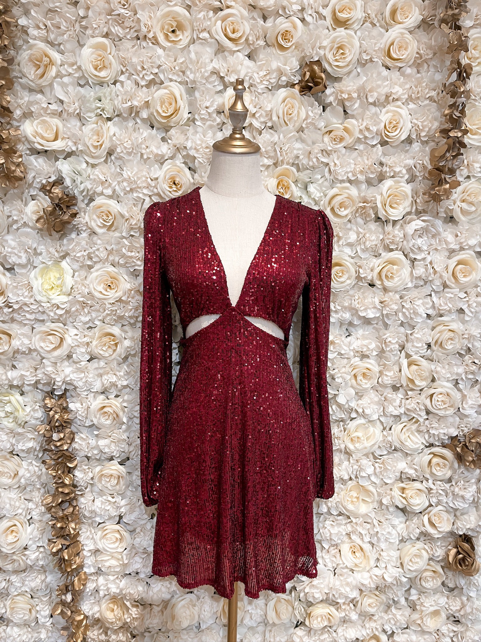 Burgundy sequin mini dress with cutouts and puff sleeves, displayed on a mannequin against a floral backdrop.