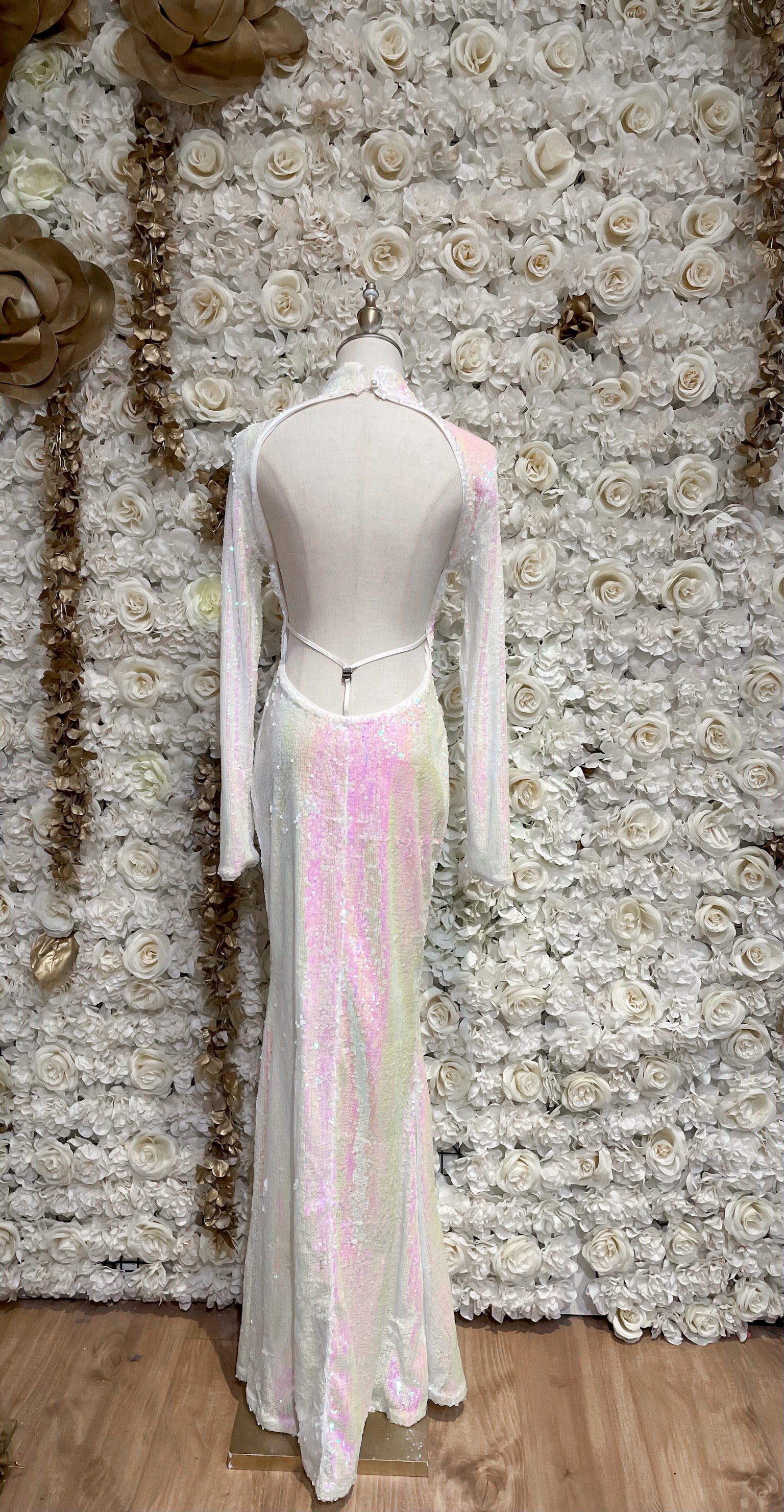 A white backless maxi dress with sequin open back, displayed on a mannequin against a floral wall backdrop.