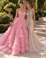 Two women wearing formal evening gowns pose outdoors on stone steps with greenery in the background. The woman on the left wears a strapless pink gown with a structured corset bodice and a very full layered tulle skirt.