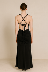 Model wearing a floor-length maxi dress with a plunging neckline, gathered detailing at the waist, and an open crisscross back with tie closure.