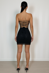 Strapless black mini dress with a sequin-embellished grid pattern, fitted silhouette, fringe-trim hemline, and an open lace-up bac