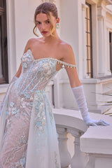 A woman models an elegant off-the-shoulder gown with intricate floral embroidery and beadwork in pale blue, white, and soft pink. She is wearing long white opera gloves and standing on a white balcony with classical columns in the background.