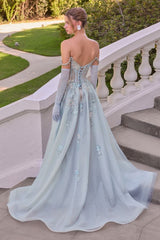 A woman wearing a formal off-the-shoulder gown in a pale icy blue tulle fabric, standing on outdoor stone steps with a garden and white balustrade behind her. The dress has long sheer gloves, a lace-up open back, and floral embroidery.