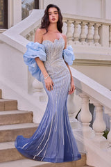 A woman models a strapless light blue mermaid gown with voluminous puff sleeves, a heavily embellished beaded bodice, and vertical silver bead lines down the skirt, posed on an outdoor stone staircase.