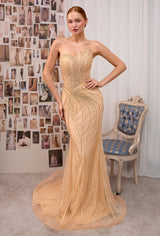 A woman models a strapless champagne beige evening gown with intricate gold beaded vertical stripes and a sweetheart neckline. She is standing in a room with a collage of fashion photos on the wall and a blue-accented vintage chair to her right.