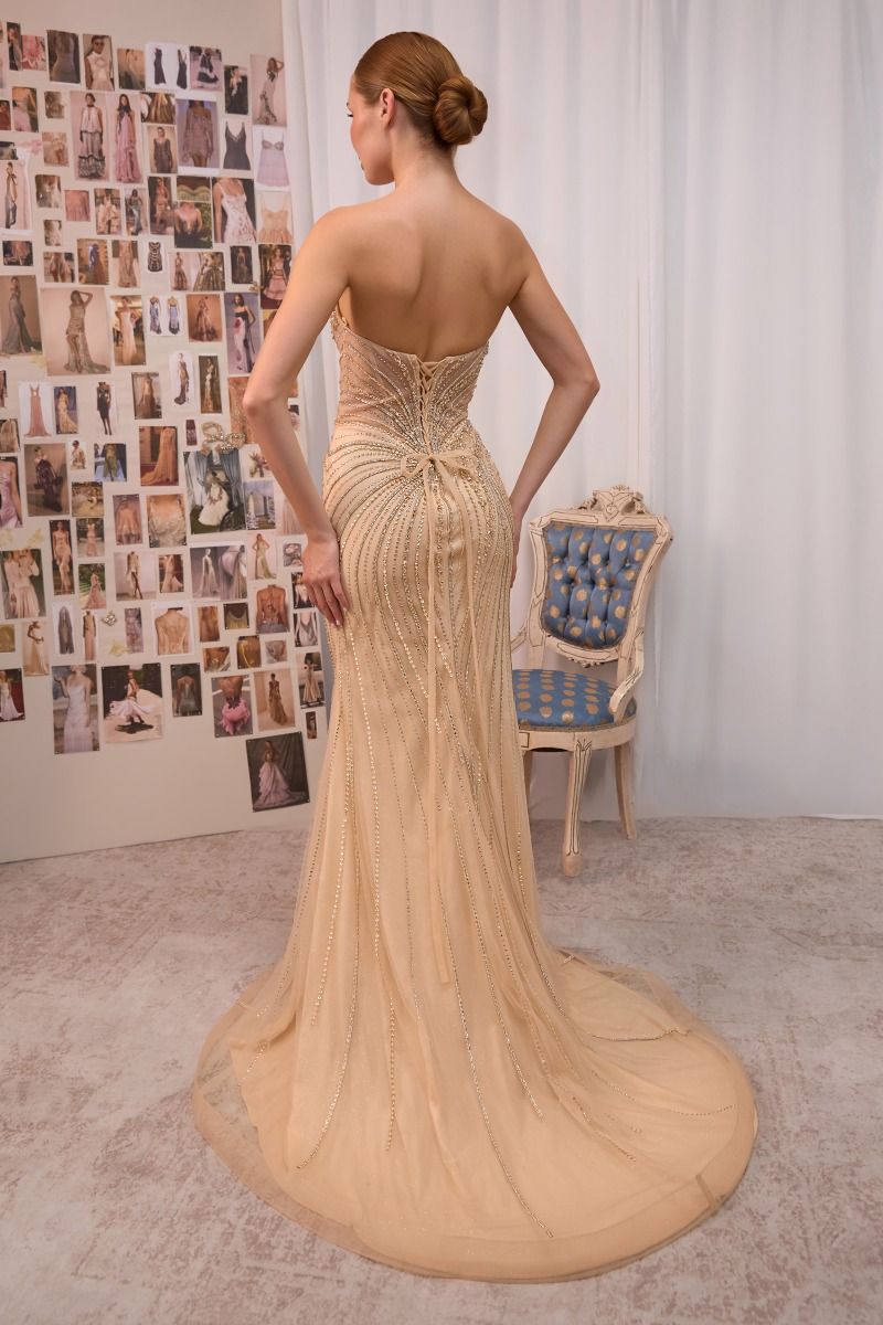 A woman stands with her back to the camera wearing a strapless champagne beige evening gown with a structured corset-style bodice and a long sheer train. The setting is an indoor studio with a white curtain backdrop, a blue upholstered chair, and a wall covered in fashion photos.