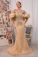A woman models a strapless champagne beige evening gown with dramatic ruffled puff sleeves, a heavily embellished beaded bodice, and a long sheer overskirt train, set against a studio backdrop with a collage board and a blue-accented chair.