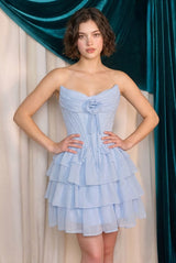 Woman wearing a light blue strapless dress with ruffled layers against a teal curtain background