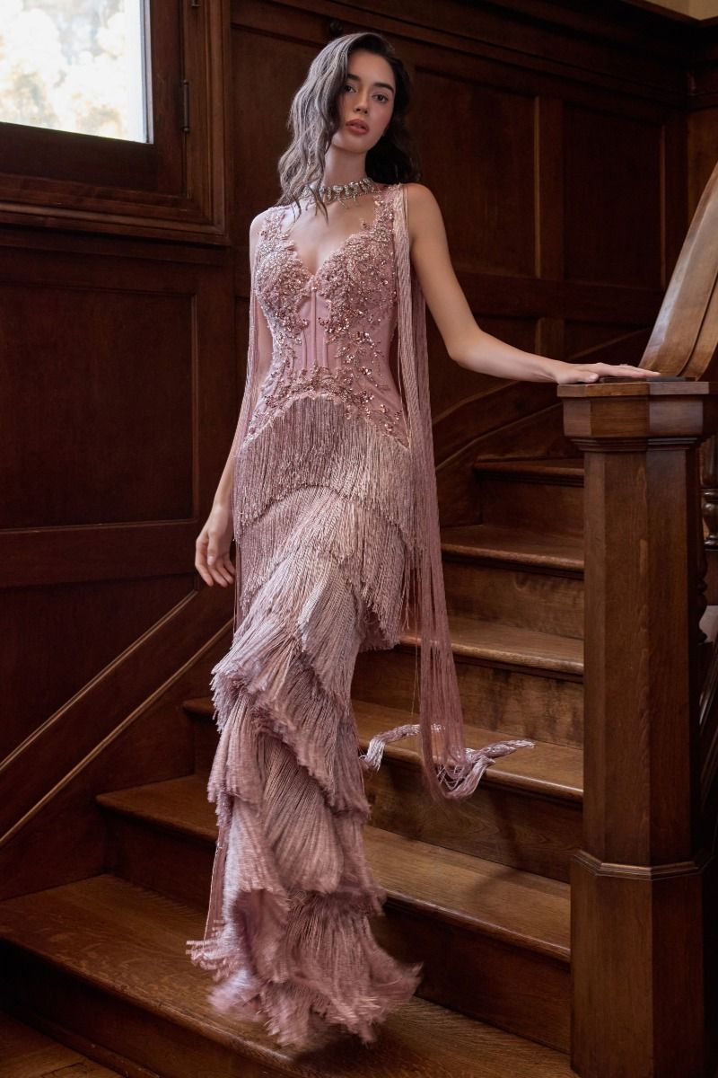 A woman wearing a sleeveless dusty pink gown with a corset bodice, fringe overlay, and layered ruffle skirt, posing on a wooden staircase in a dimly lit room.