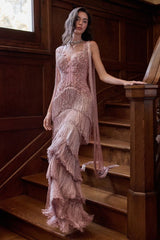 A woman wearing a sleeveless dusty pink gown with a corset bodice, fringe overlay, and layered ruffle skirt, posing on a wooden staircase in a dimly lit room.