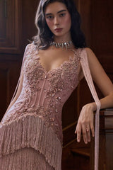 A woman wearing a sleeveless dusty pink evening gown with intricate beadwork, sequins, and a long fringe hem, posed against a dark wood interior background.