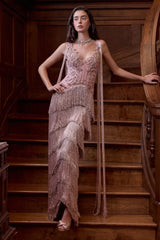 A woman stands on wooden stairs wearing a sleeveless, heavily embellished floor-length gown in a dusty blush pink tone with cascading fringe and long draped side details. The setting is a dark wood interior staircase.