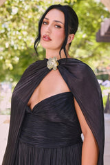 A woman wearing a black pleated dress with a draped cape-style overlay and a large decorative gold flower accent at the neckline, photographed outdoors with blurred green foliage in the background.