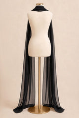 A long sheer black cape draped over a cream dress form mannequin on a gold stand, set against a solid beige background.