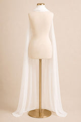 A long, flowing white bridal veil draped over a cream-colored dress form mannequin on a gold stand, set against a solid beige background.