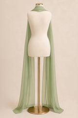 A sage green sheer chiffon cape draped over a cream-colored dress form mannequin on a gold stand, set against a solid warm beige background.