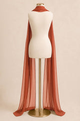 A rust-orange sheer cape scarf draped over a cream-colored mannequin torso on a gold stand, set against a solid beige background.