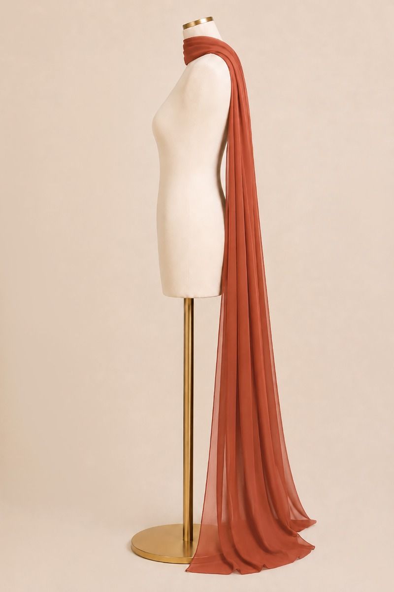 A long, semi-sheer rust-colored scarf draped over a cream dress form mannequin on a gold stand, set against a solid beige background.