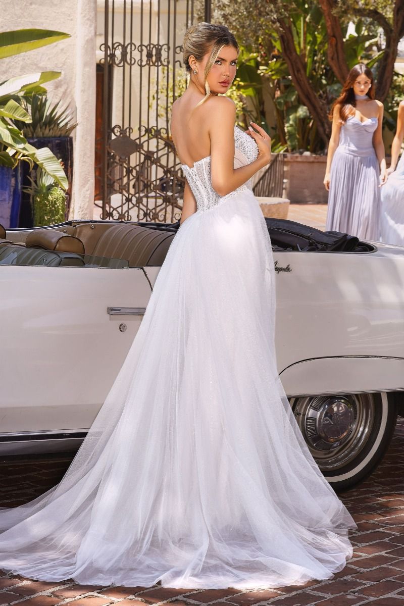 Woman in a white wedding dress standing next to a vintage car with another woman in a white dress in the background.