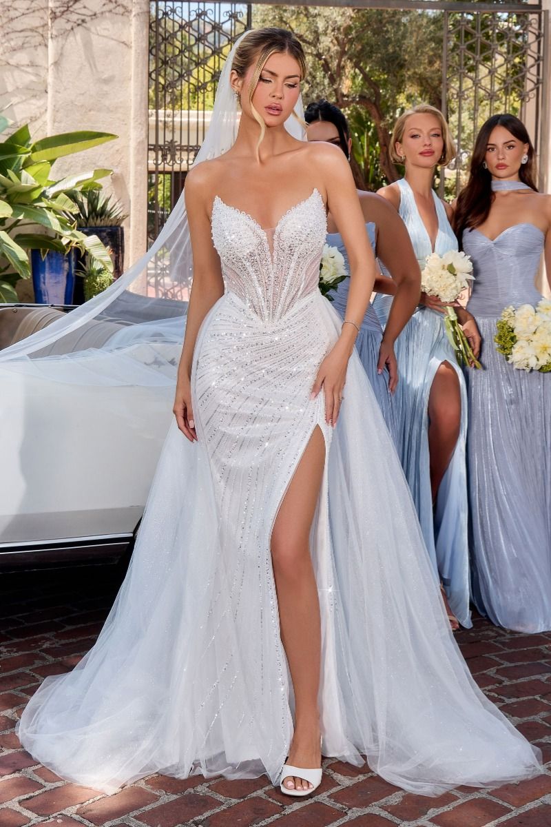 Woman in a white wedding dress with a high slit, standing outdoors with other women in light blue dresses.