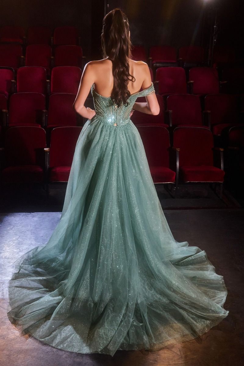 A woman stands with her back to the camera wearing a floor-length off-the-shoulder gown in a dimly lit auditorium with rows of red seats. The dress is a soft sage green with shimmering embellishments and a full tulle skirt.