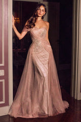 A woman models a strapless floor-length evening gown in a dusty blush pink tulle fabric with intricate silver sequin and bead embellishments. She is posed in a doorway with a dark interior background.