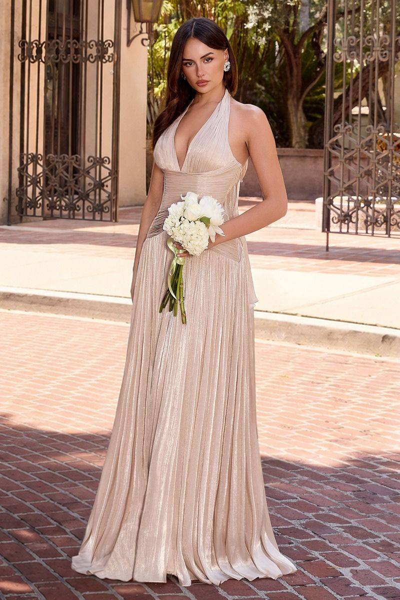 A woman stands outdoors on a brick walkway wearing a sleeveless, halter-neck, floor-length champagne pleated gown and holding a bouquet of white flowers.